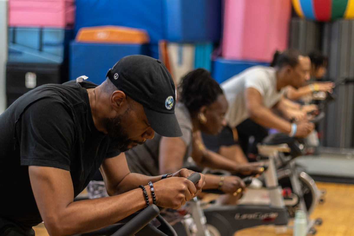 A group leans over the handlebars of their exercise bikes while participating in a workout class.