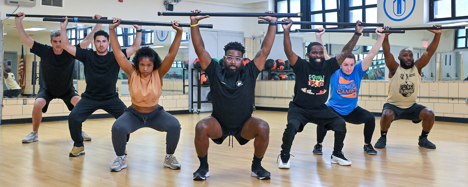 With exercise bars over their heads, a group of people led by a fitness instructor squat and look into the camera.