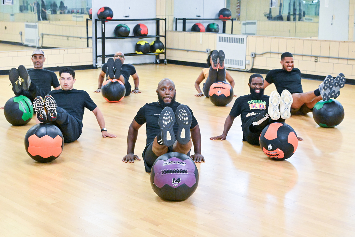 A group led by a fitness instructor hold their legs up and over an exercise ball while seated on the floor.