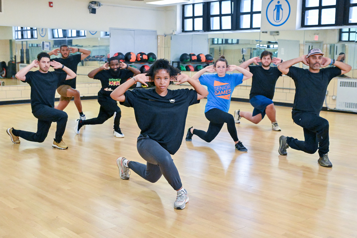 A group led by a fitness instructor cross their legs in a crouching lunge with their hands behind their heads.